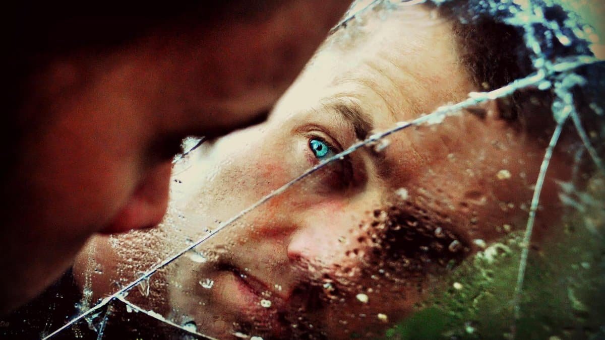 A man with green eyes peers into a cracked, wet mirror, conveying solitude and thought.