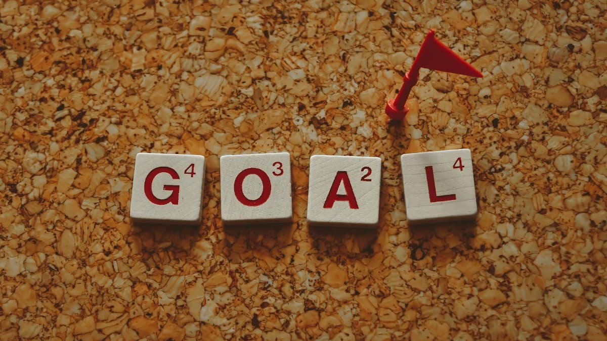 Scrabble tiles forming the word 'goal' with a small red flag on a cork board background.