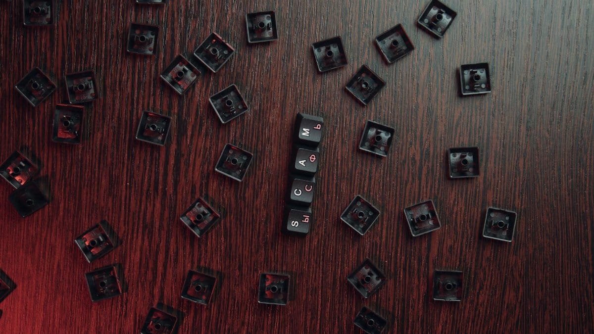 Aerial view of scattered keyboard keycaps on a wooden surface with 'SCAM' spelled out.