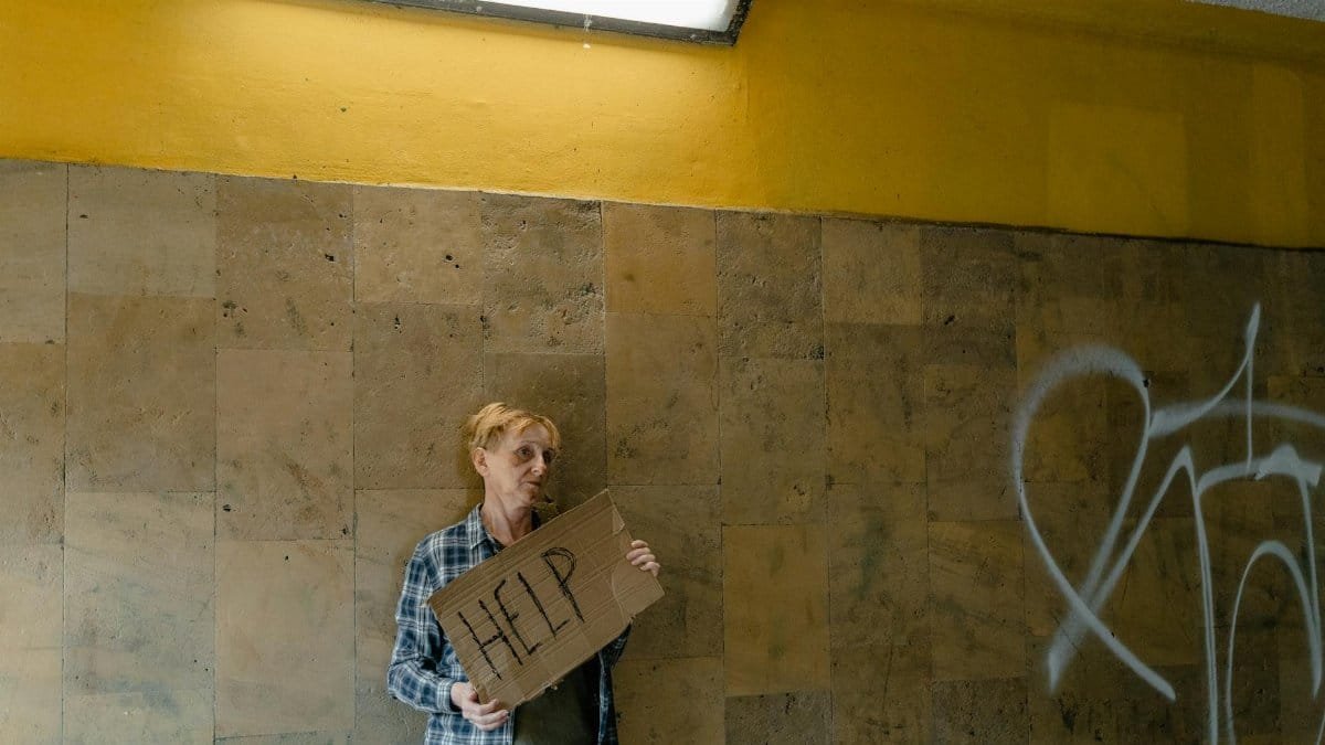 A homeless person holding a cardboard sign asking for help in an urban environment.
