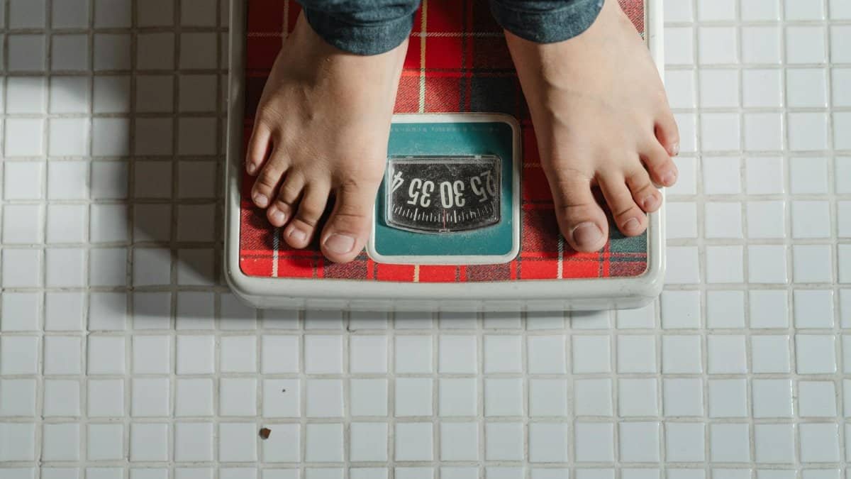 From above crop anonymous barefoot child in jeans standing on weigh scales on tiled floor of bathroom
