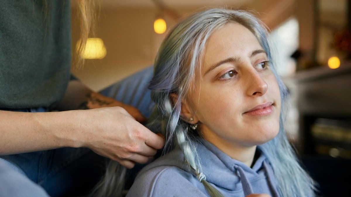 A young woman with gray hair getting a braid done indoors with a gentle smile.