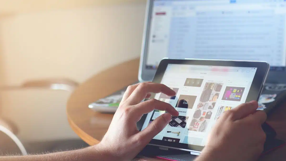 Hands using a tablet beside a laptop in a home office setting, showcasing online browsing.