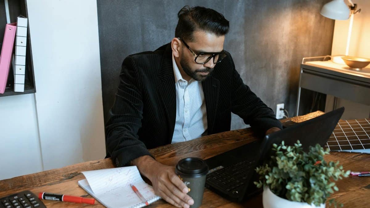 Man concentrating on laptop in a modern home office setting, coffee in hand.