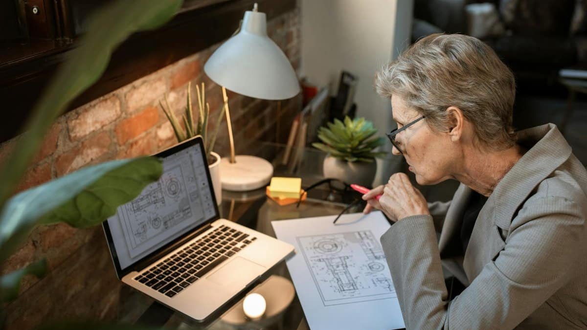 A senior engineer analyzes architectural plans on a laptop in a cozy home office.