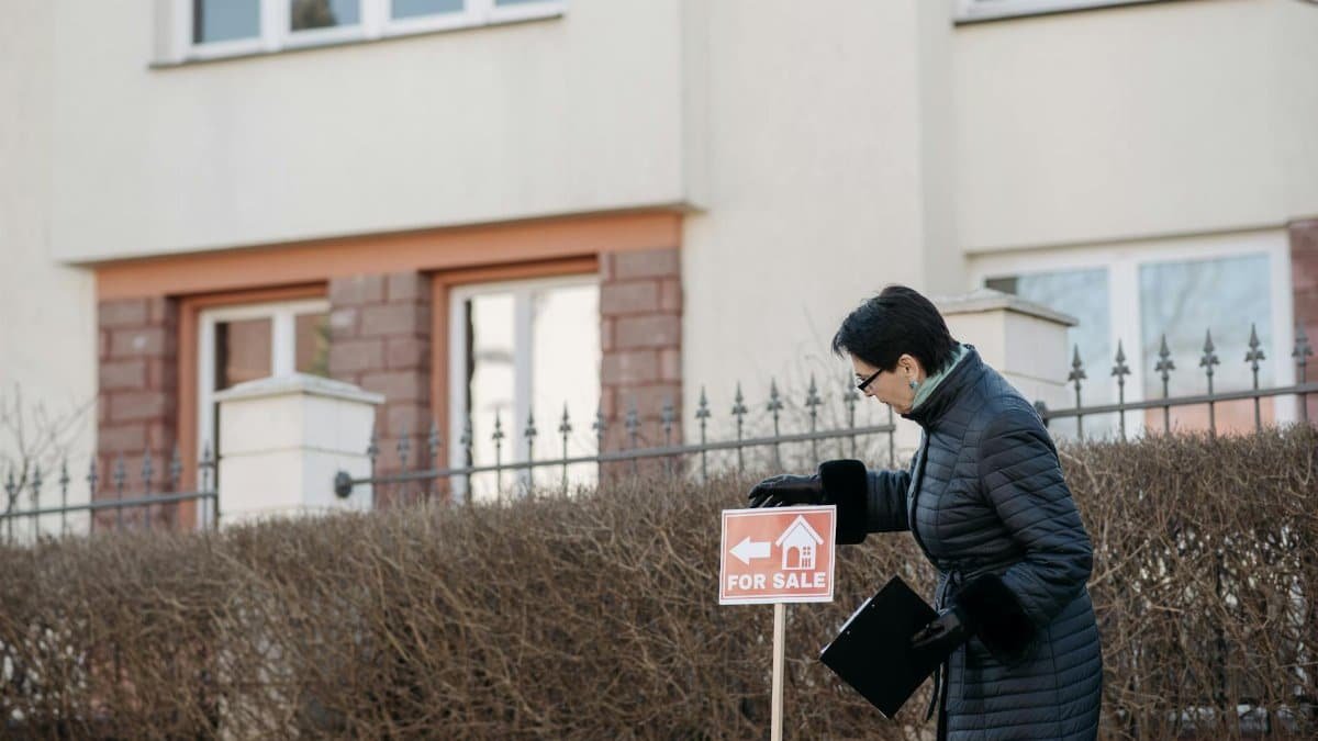 Real estate agent in a black coat placing a 'For Sale' sign in front of a house.