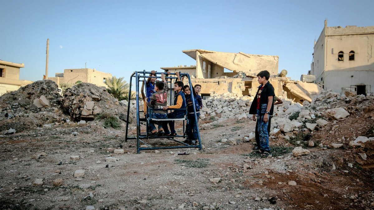 Children play on a swing set in a war-torn area of Idlib, Syria, showcasing resilience.