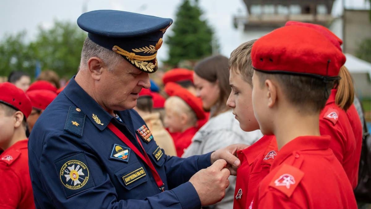 A Russian officer in uniform pinning a badge on a young recruit during an outdoor military ceremony.