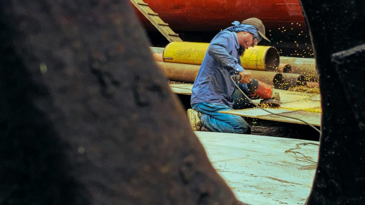 Man working in shipyard, using power tool to cut metal and create sparks.
