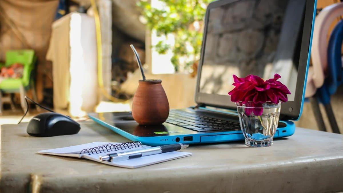 A serene outdoor workspace featuring a laptop, notebook, mug, and flowers on a desk.