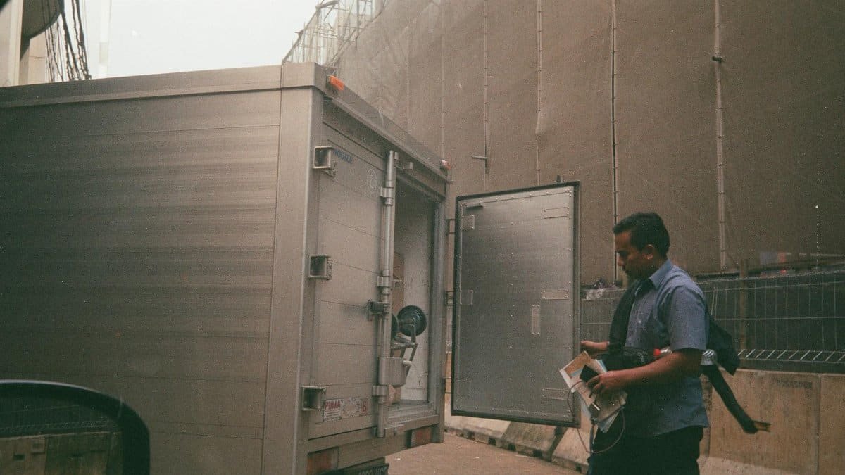 A logistics worker in a blue shirt checking a cargo truck on a city street. Industrial background suggests transportation and delivery.