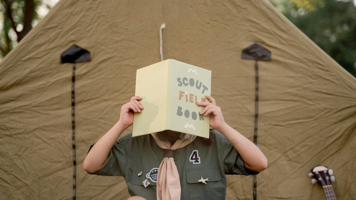 A scout child reads a field book outside a tent, embracing outdoor adventure.
