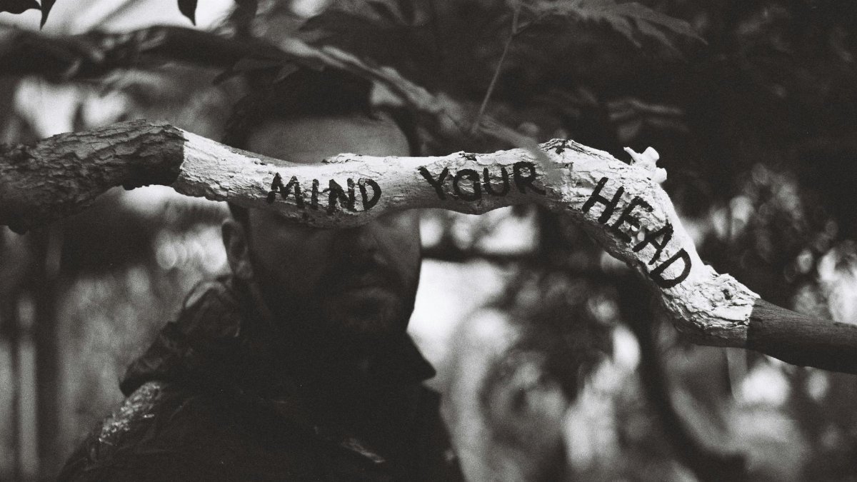 Black and white photo of a 'Mind Your Head' sign on a branch with a man in the background.