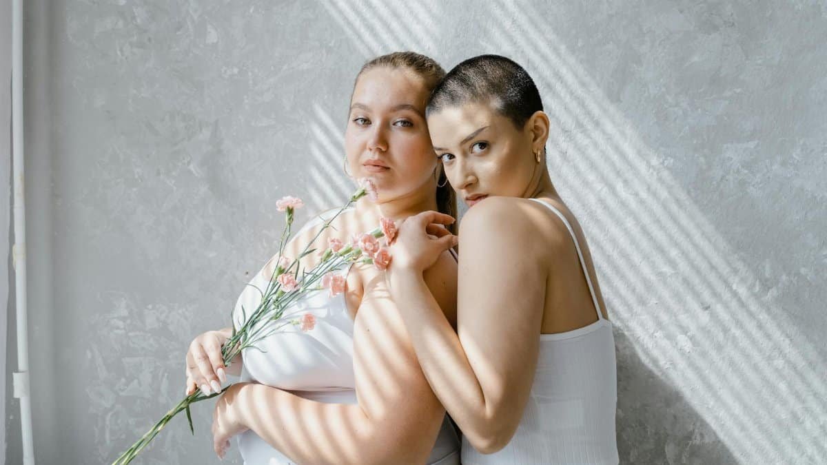 Two women confidently pose with flowers in an indoor setting, exuding self-esteem and empowerment.