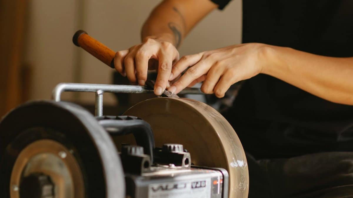 Unrecognizable male master with tattoo sharpening metal detail on grinding wheel while sitting at instrument in professional workshop on blurred background