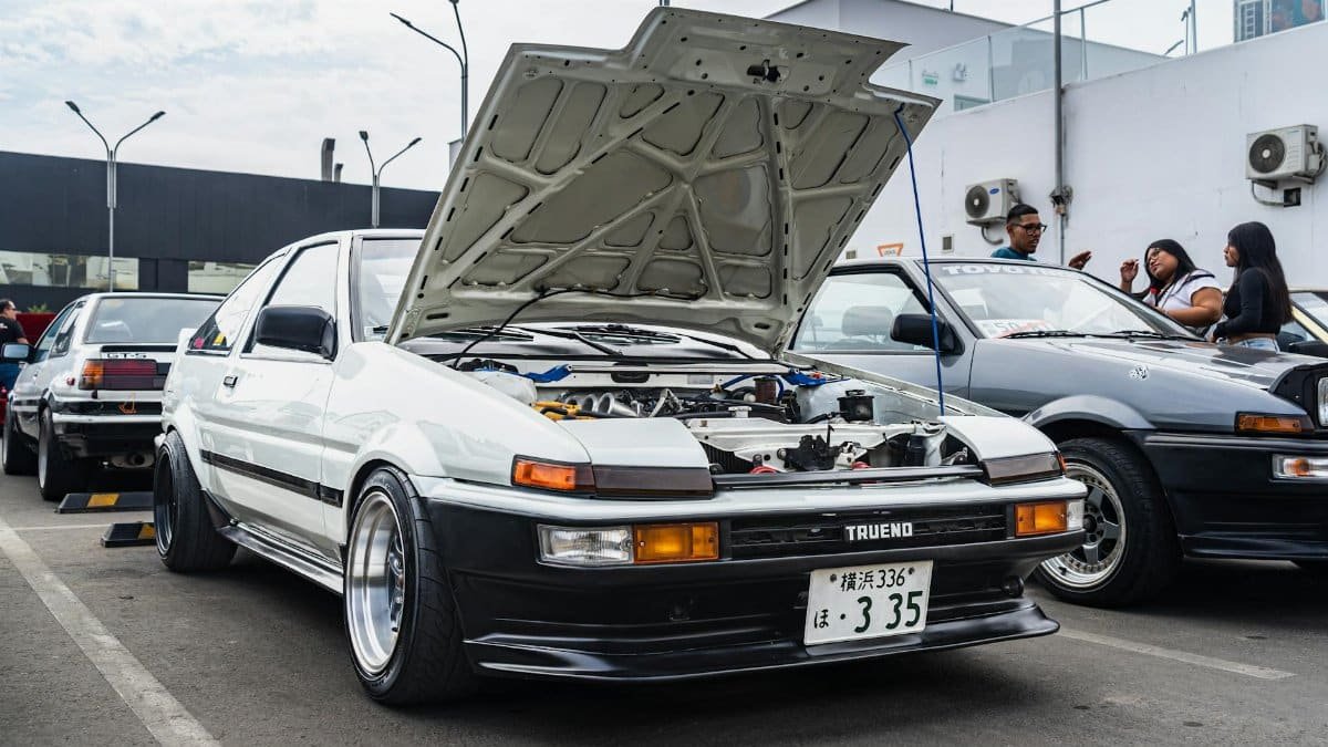 White Toyota AE86 Trueno with open hood at a city car show parking lot.