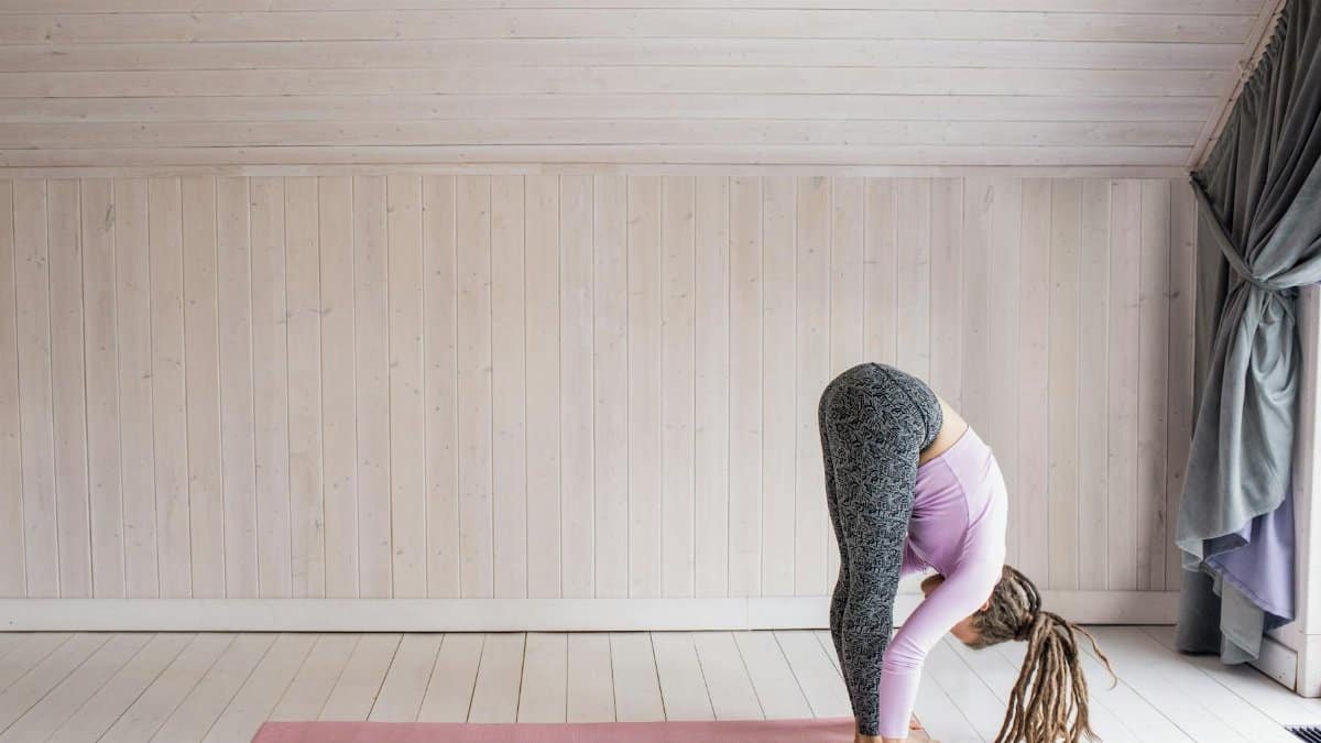 A woman doing a forward bend yoga pose on a pink mat in a serene indoor setting.