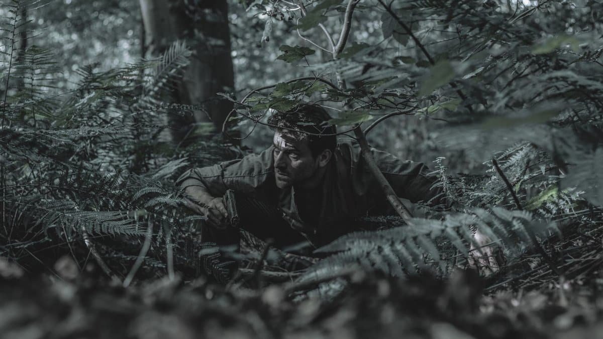 A man crouches in dense forest foliage, blending with his surroundings, evoking a sense of adventure and stealth.