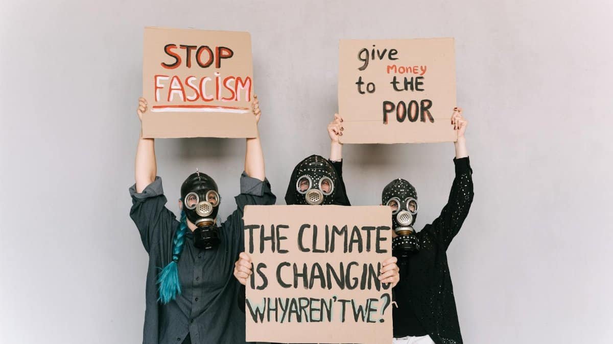 Three people wearing gas masks hold signs advocating social change and awareness.
