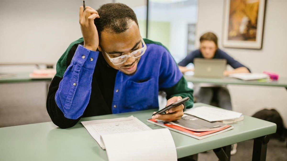 A college student studying in class using a smartphone, with papers and books on the desk.