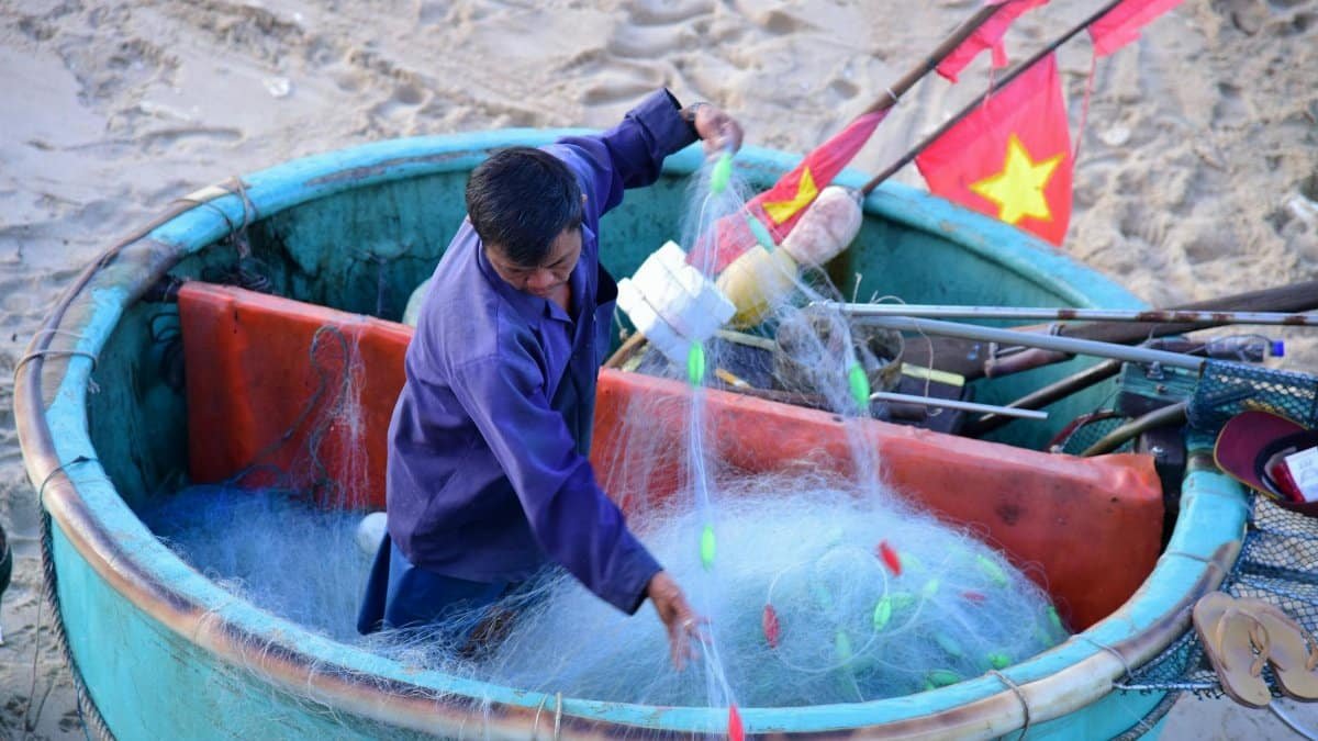 A fisherman operates a traditional Vietnamese round boat, preparing fishing nets on a sandy beach.
