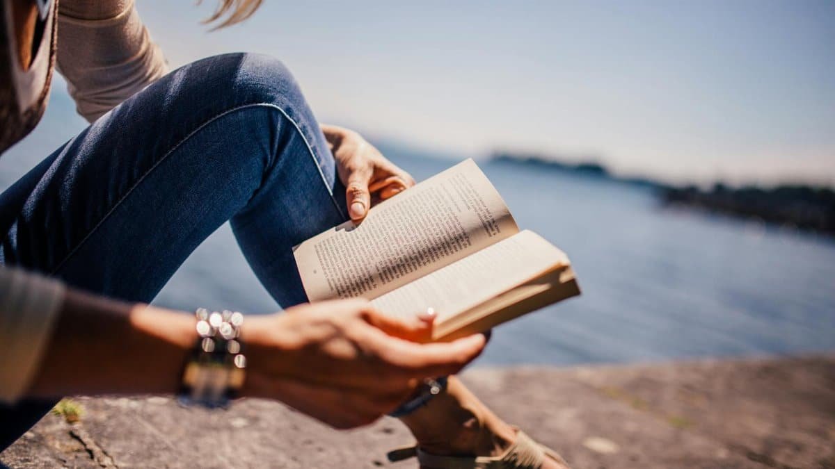 Serene image of a young woman reading by the lake under a clear blue sky.