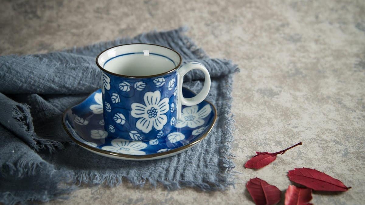 Elegant floral porcelain coffee mug on a blue cloth with red leaves and textured background.