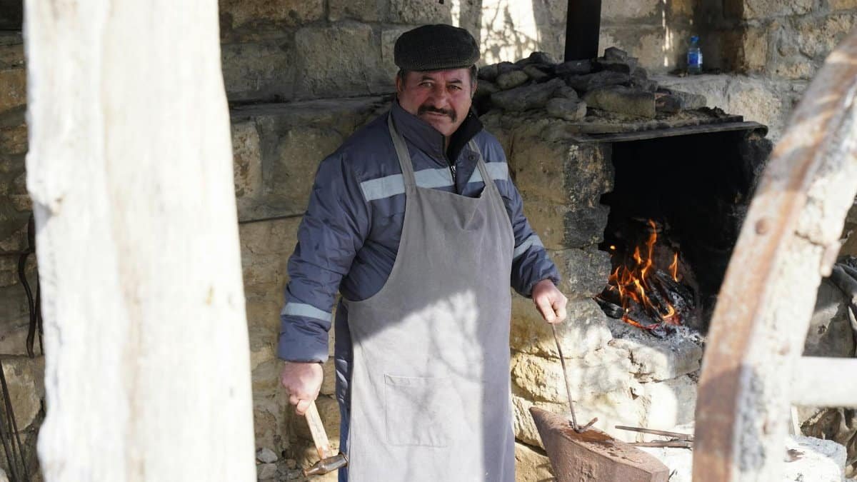 A blacksmith works at a traditional forge outdoors, wearing an apron and holding tools.