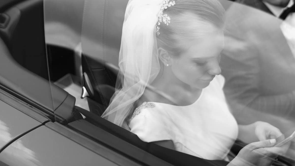 From above black and white of woman in veil text messaging on cellphone near crop anonymous man in automobile on wedding day