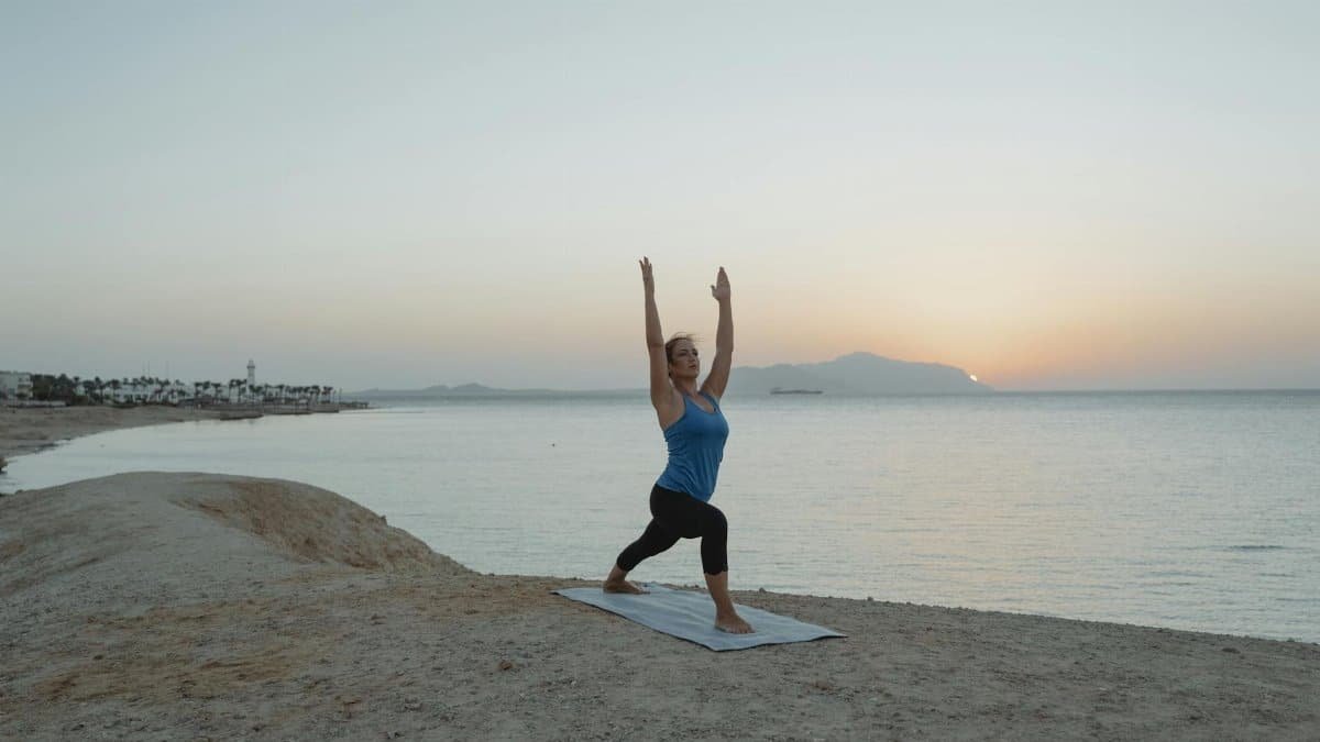 A woman practices Warrior 1 yoga pose on a beach at sunset, embracing tranquility and balance.