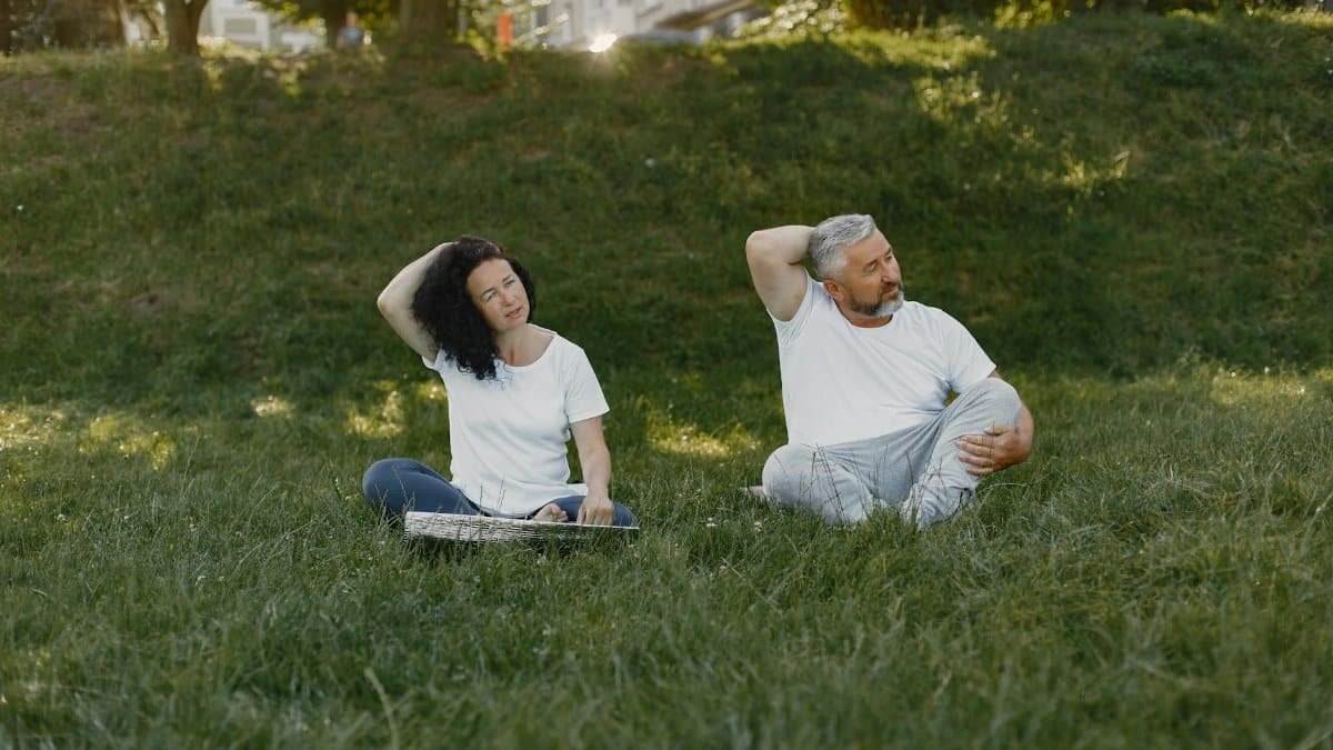 Couple doing neck stretches outdoors, promoting a healthy lifestyle in a sunny park setting.