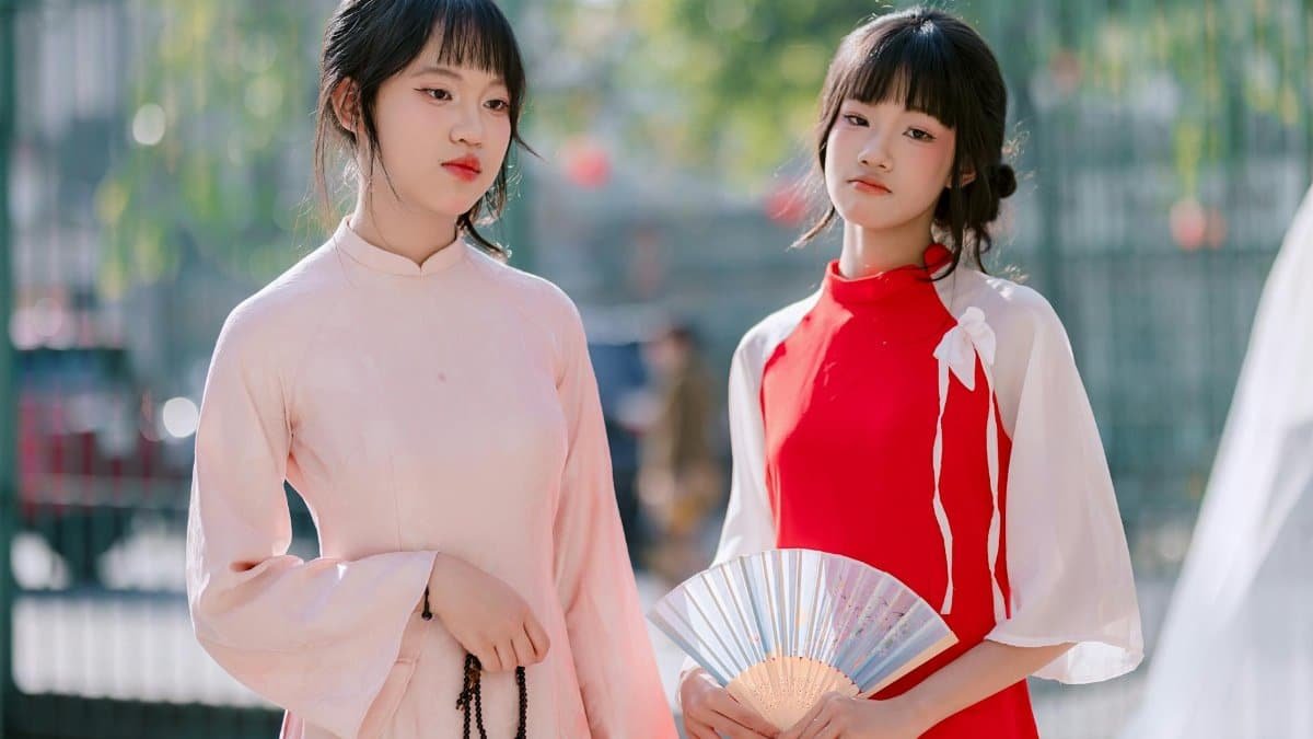 Two women in traditional dresses posing with a fan outdoors in a serene setting.