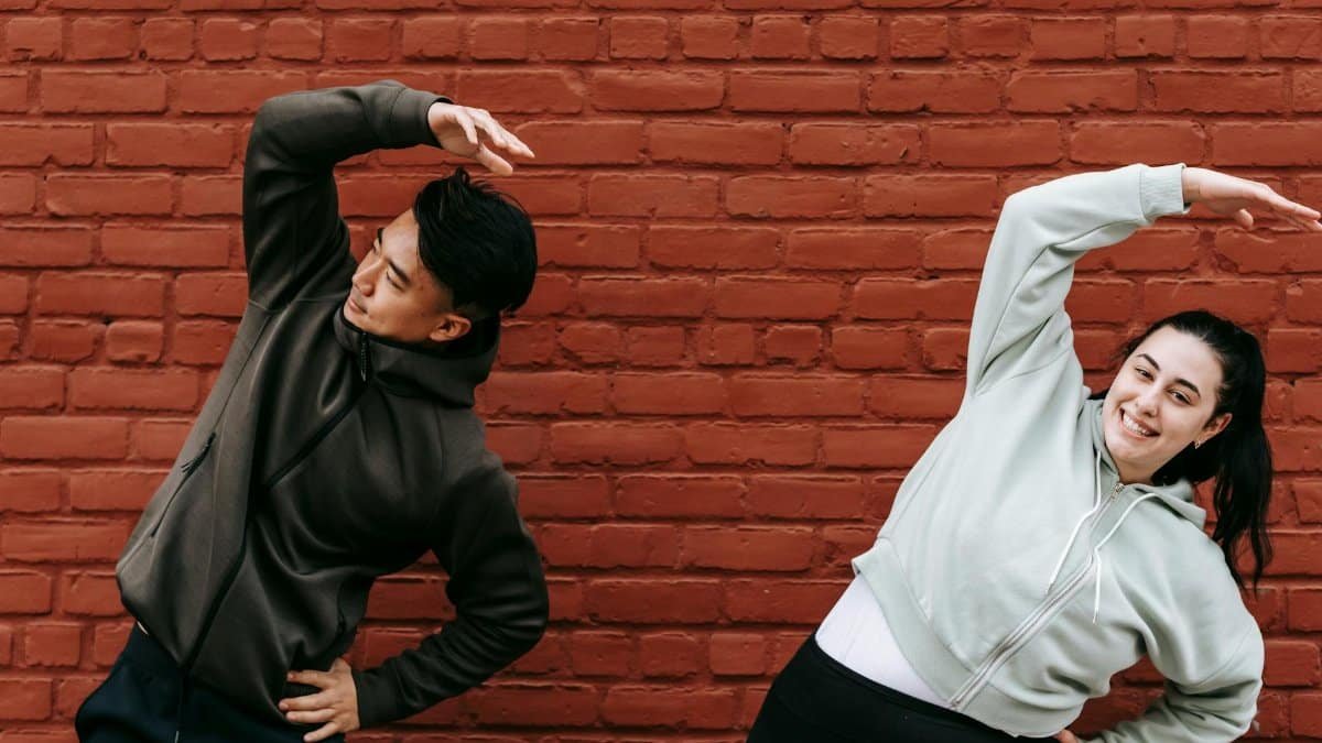 Positive young plus size female and fit Asian male trainer standing near brick wall and doing Overhead Side Reach Stretch exercise before workout on street
