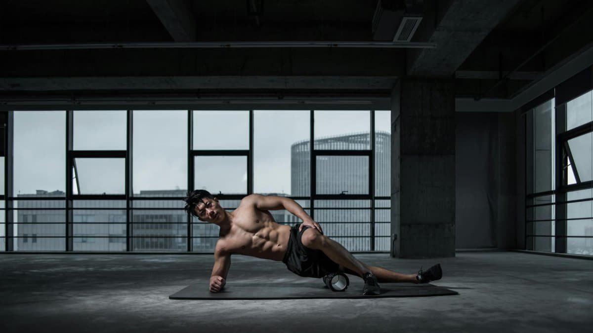 Muscular man performing yoga stretch indoors on a mat with urban window view in the background.