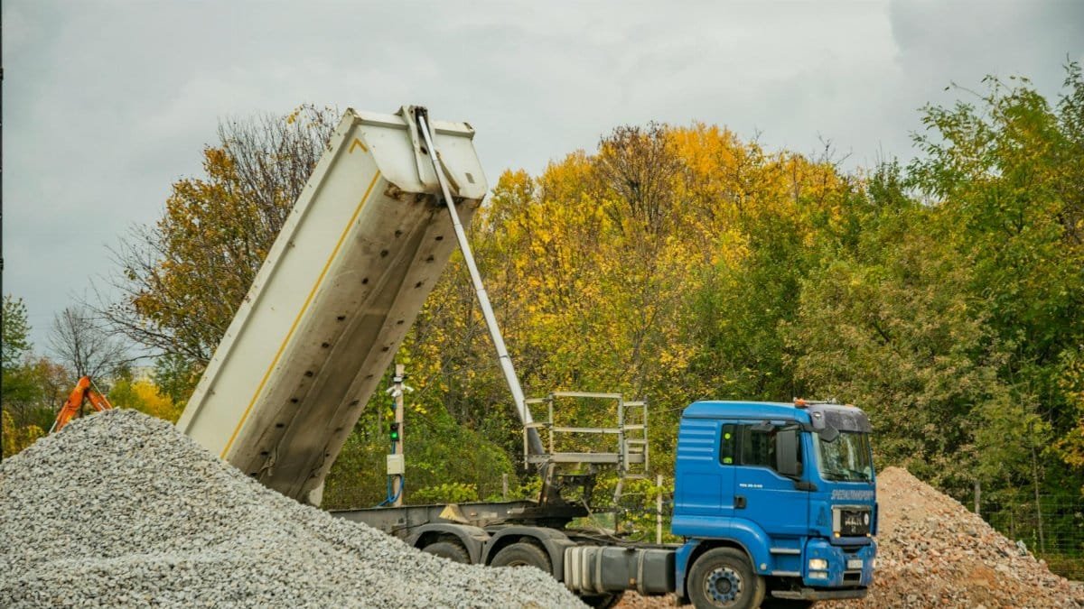 Blue dump truck unloading gravel at an outdoor construction site surrounded by autumn trees.