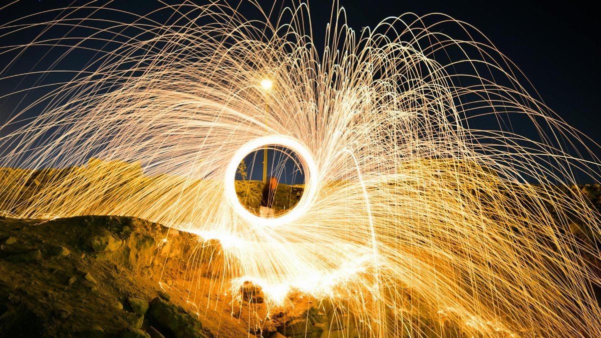 Captivating long-exposure shot of steel wool spinning, creating vivid sparks against a rocky terrain at night.