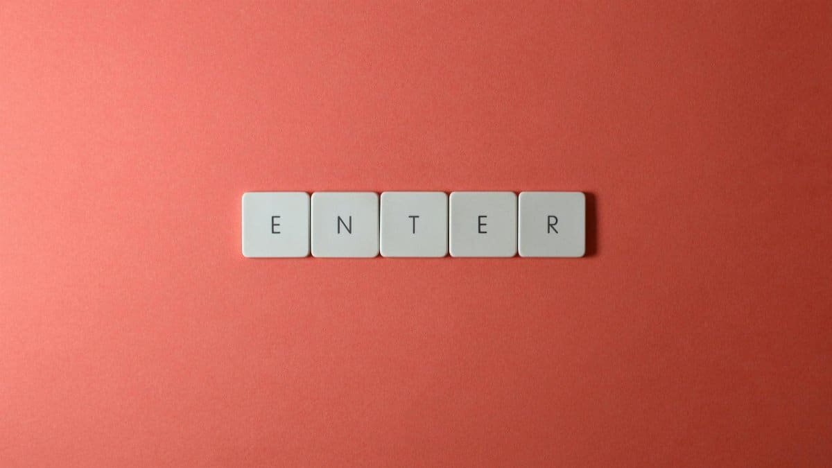 Close-up of five keyboard letter tiles spelling 'ENTER', placed on a coral backdrop.