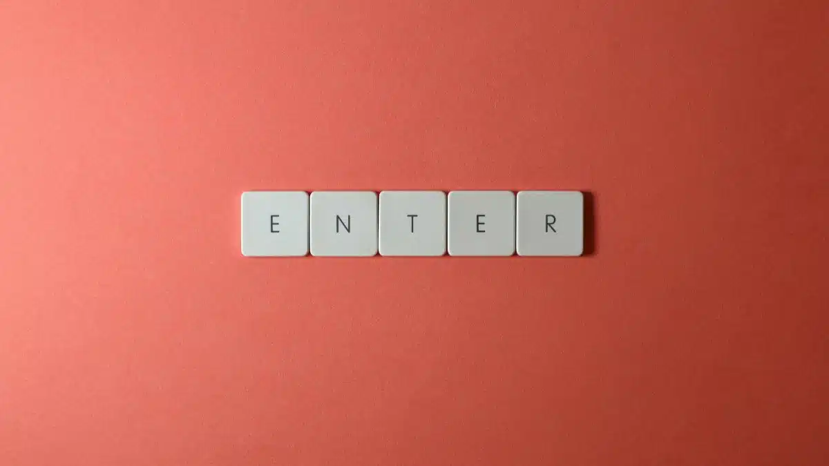 Close-up of five keyboard letter tiles spelling 'ENTER', placed on a coral backdrop.