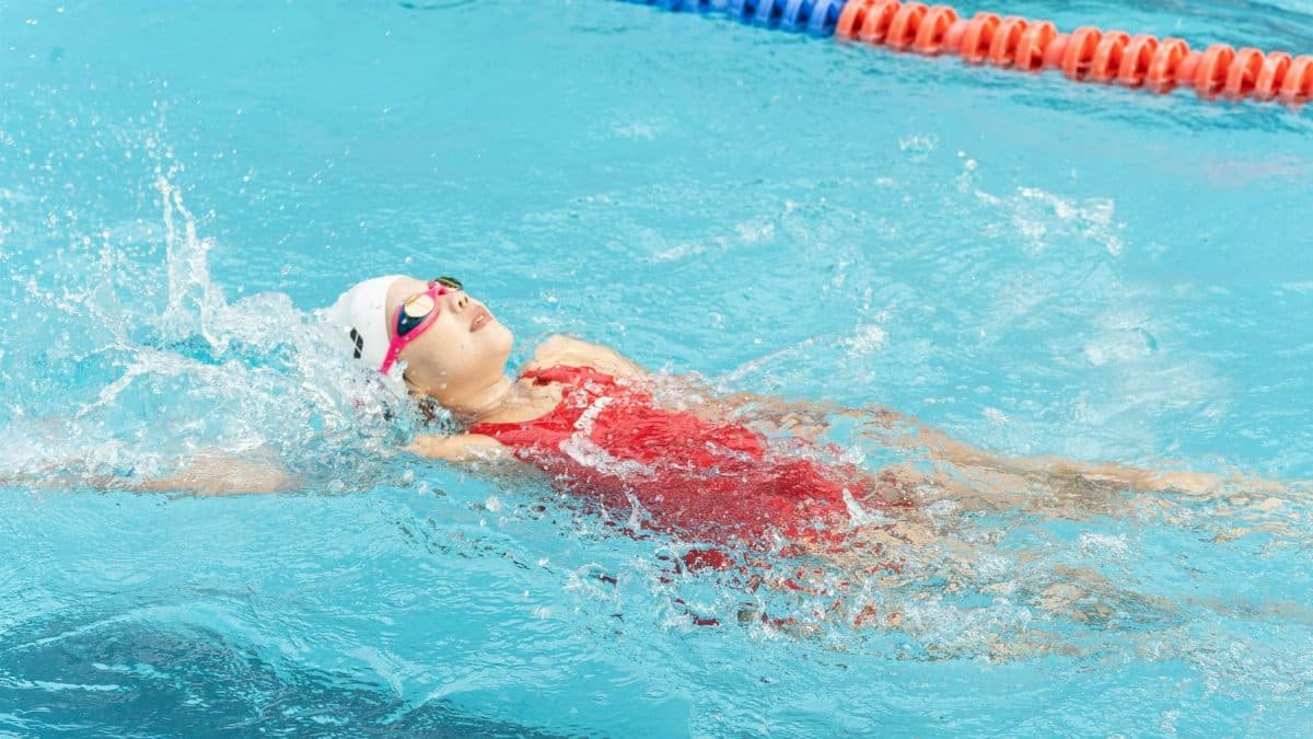 Young swimmer practicing backstroke in a swimming pool, wearing goggles and swim cap.