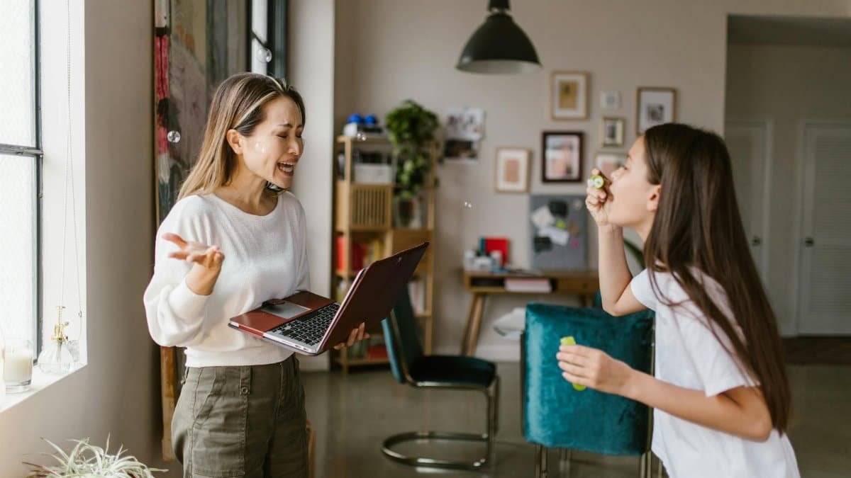 A mother works remotely while her daughter plays with bubbles in a cozy home setting.