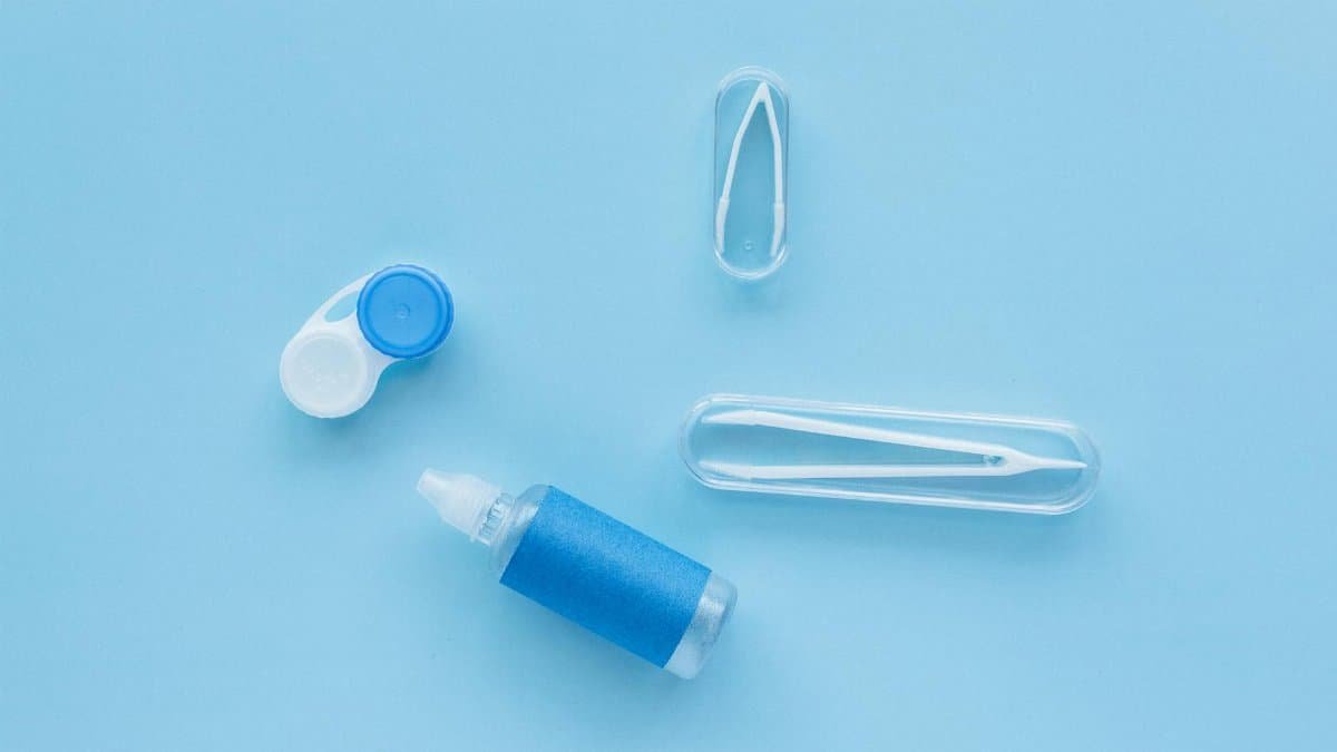 Flat lay of contact lens case, tweezers, and solution bottle on blue background.
