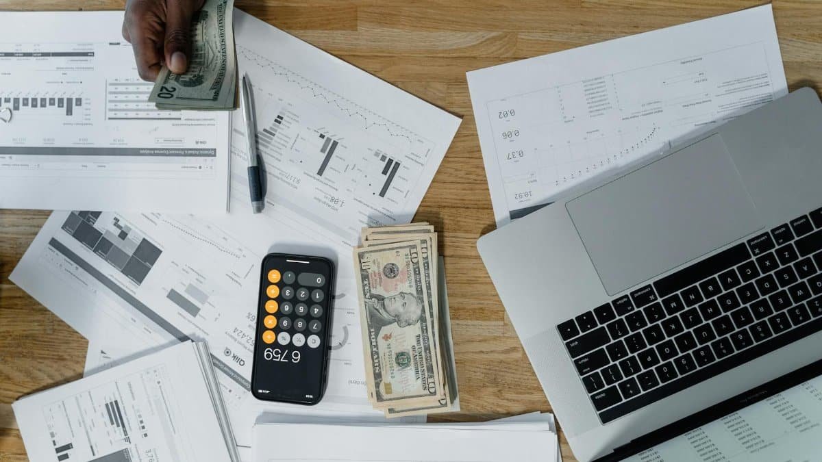 Overhead view of a busy workspace with cash, financial documents, and a laptop.