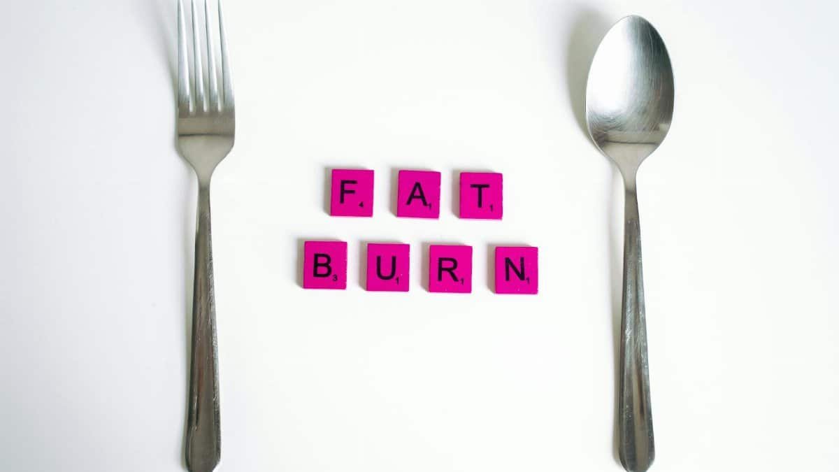 Conceptual art featuring the words 'Fat Burn' using Scrabble tiles between a fork and spoon on a white background.