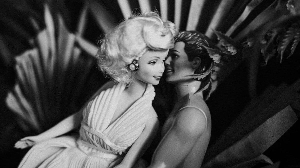 Vintage-style black and white photo of Barbie and Ken dolls in a romantic embrace amidst foliage.