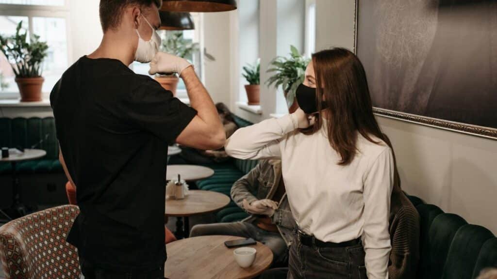 Two friends greeting with elbow bump in a café, wearing masks for safety.