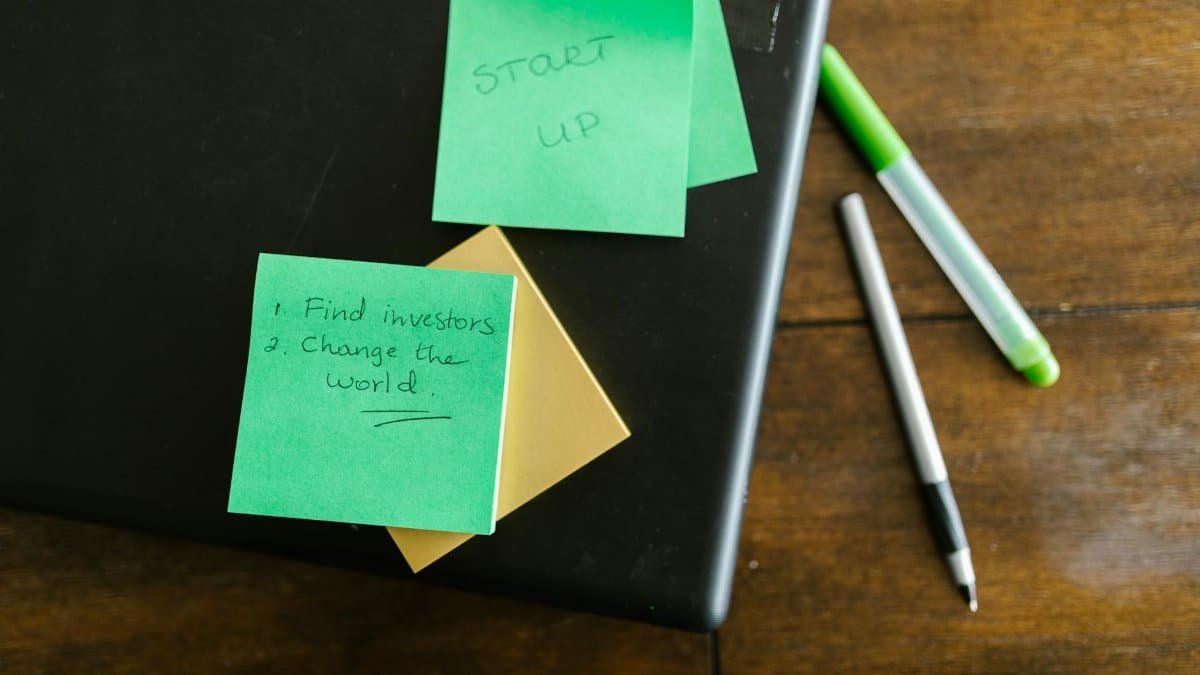 Green sticky notes with startup goals on a wooden desk with pens.