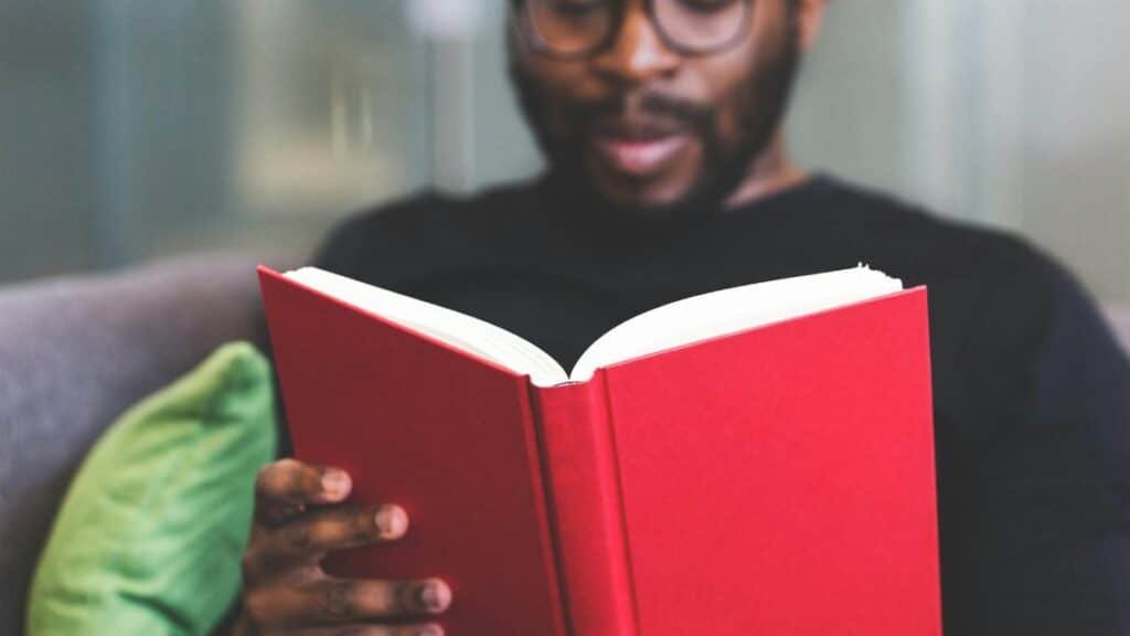 Focused young man reading a red book indoors, showcasing learning and relaxation.