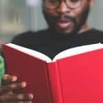 Focused young man reading a red book indoors, showcasing learning and relaxation.