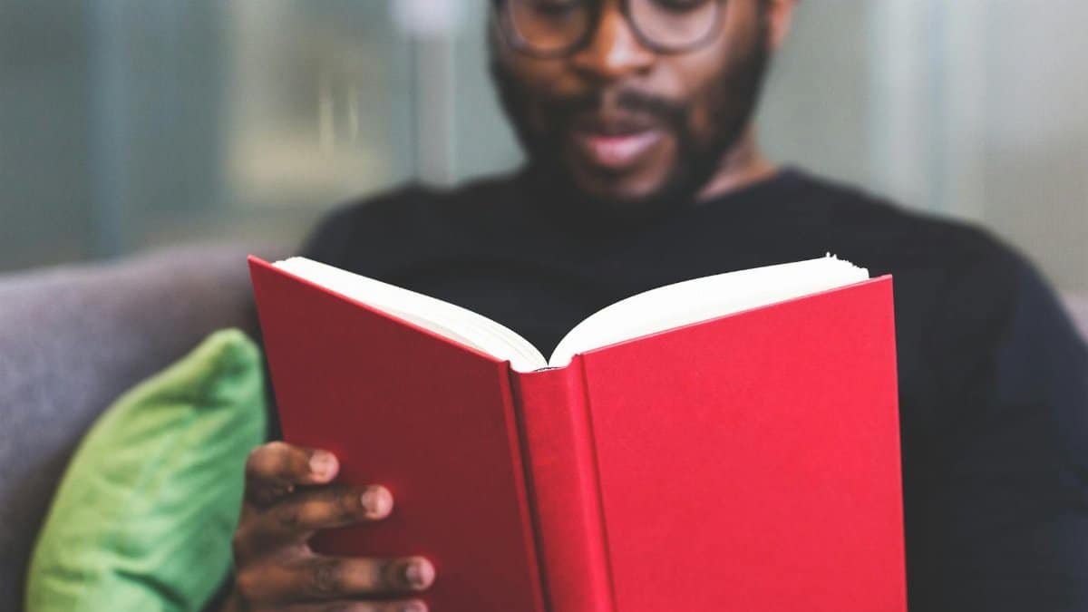 Focused young man reading a red book indoors, showcasing learning and relaxation.