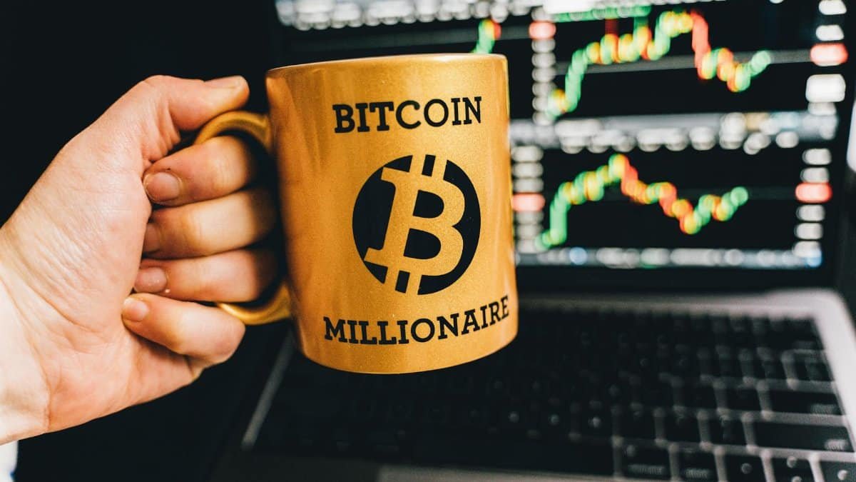 Golden mug with 'Bitcoin Millionaire' text in front of stock market charts on laptop screen.